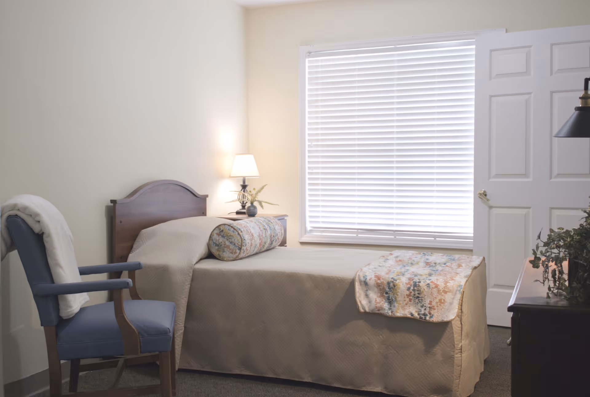 A simple bedroom with a single bed covered in a beige quilt, a floral bolster pillow, and a matching floral throw at the foot. Next to the bed is a wooden nightstand with a small lamp and a decorative plant. A blue upholstered chair with a white blanket draped over the back is positioned near the bed. A window with white blinds is behind the bed, and a white door is partially open to the right.