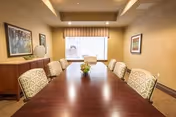 A long wooden table with upholstered chairs and a small plant centerpiece in a softly lit meeting/dining room with artwork and a window.