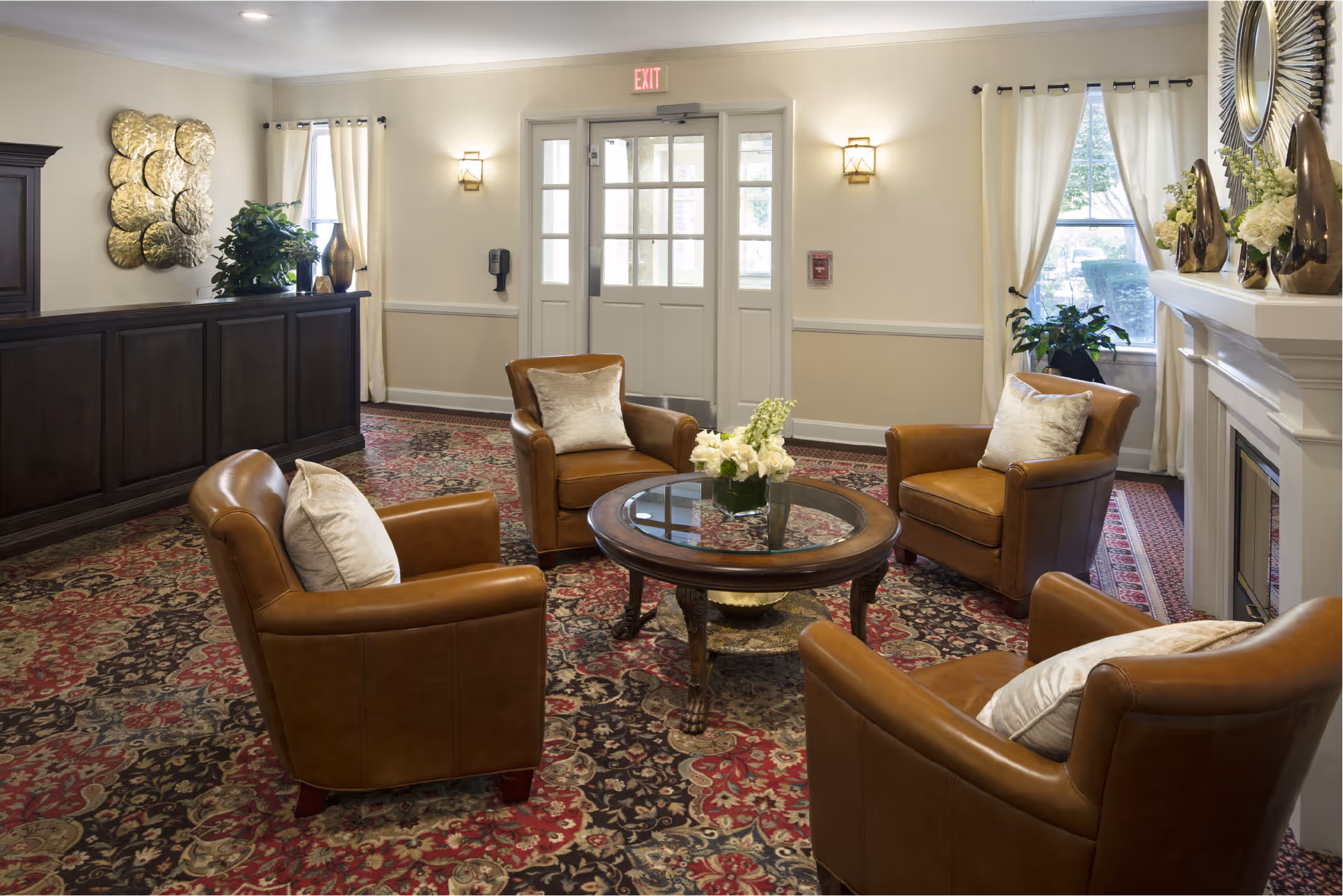 A cozy sitting area with four brown leather armchairs arranged around a round glass-top wooden coffee table with a flower arrangement. The room has a patterned carpet, cream-colored walls, a fireplace with decorative vases and flowers, and large windows with white curtains letting in natural light. There is a dark wooden reception desk on the left side and double doors with glass panels at the back.