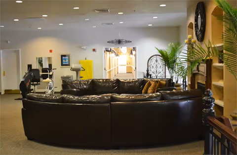 Interior view of a common area in an assisted living facility featuring a large curved dark leather sectional sofa with several cushions. Behind the sofa, there is exercise equipment and a hallway leading to other rooms. The room is decorated with plants, a large clock on the wall, and built-in shelves with decorative items.