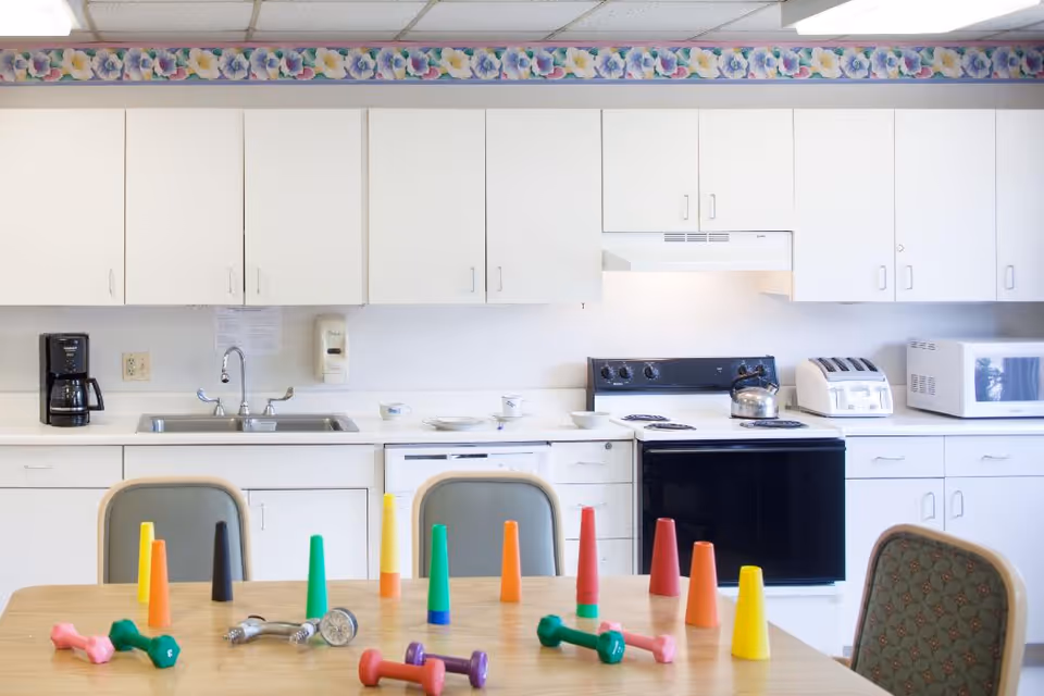 A bright kitchen area with white cabinets, a black stove, a microwave, a toaster, and a coffee maker. In the foreground, there is a table with colorful plastic cones and small dumbbells arranged on it. Two chairs are partially visible around the table. The kitchen has a floral border wallpaper near the ceiling.
