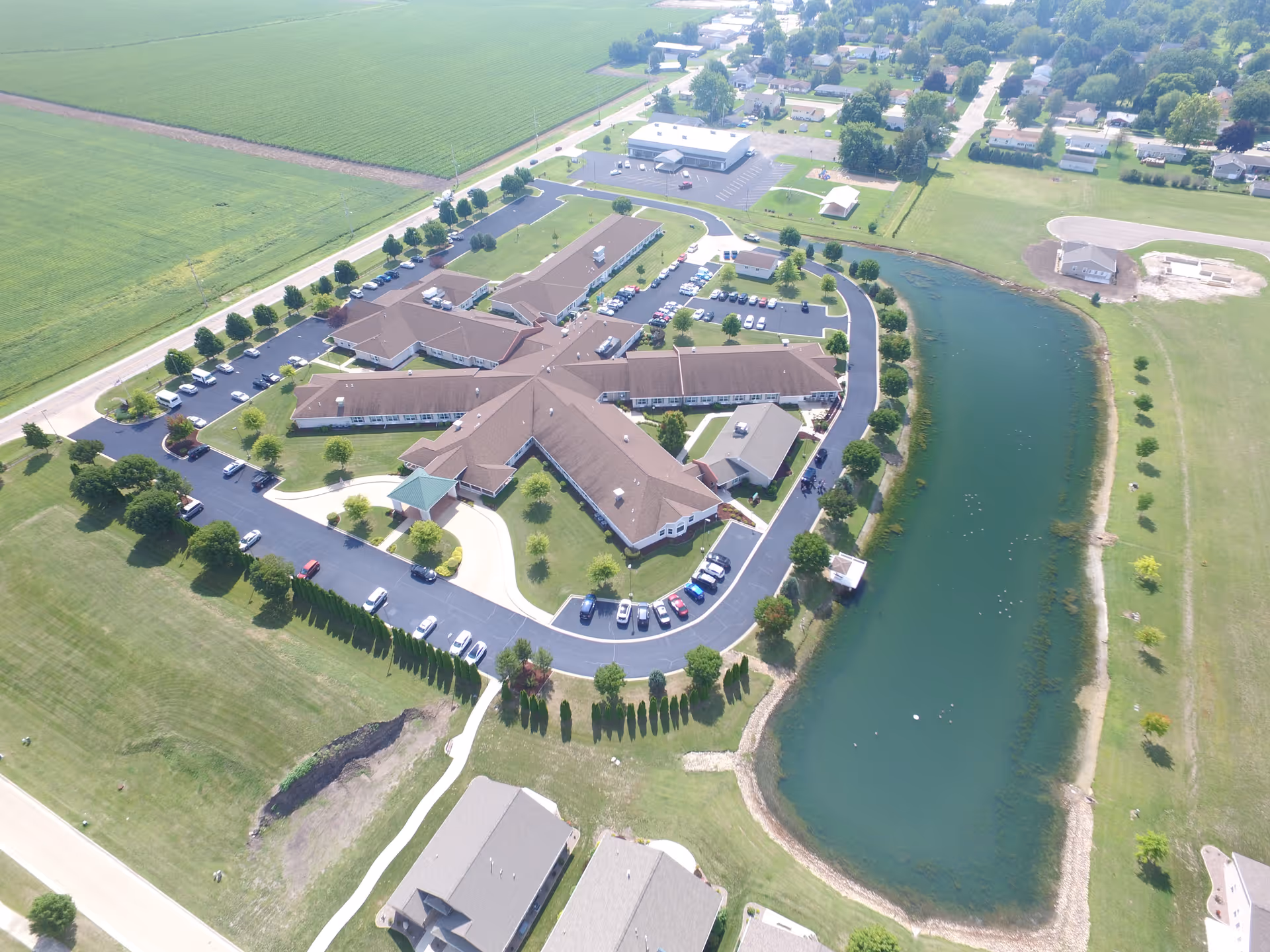 Aerial view of Liberty Village of Princeton senior living facility showing a large building complex with multiple wings, surrounded by parking lots, green lawns, trees, and a pond on the right side.
