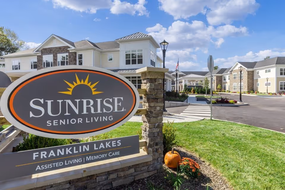 Exterior view of Sunrise Senior Living facility in Franklin Lakes with a large sign in the foreground displaying the facility name and services. The building is two stories with white and stone facade, surrounded by a well-maintained lawn and a clear blue sky with some clouds.