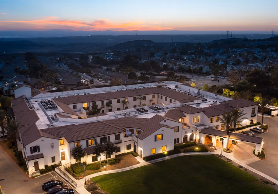Aerial dusk view of a Mediterranean-style senior living building complex with lit windows, entry canopy, and surrounding neighborhood.