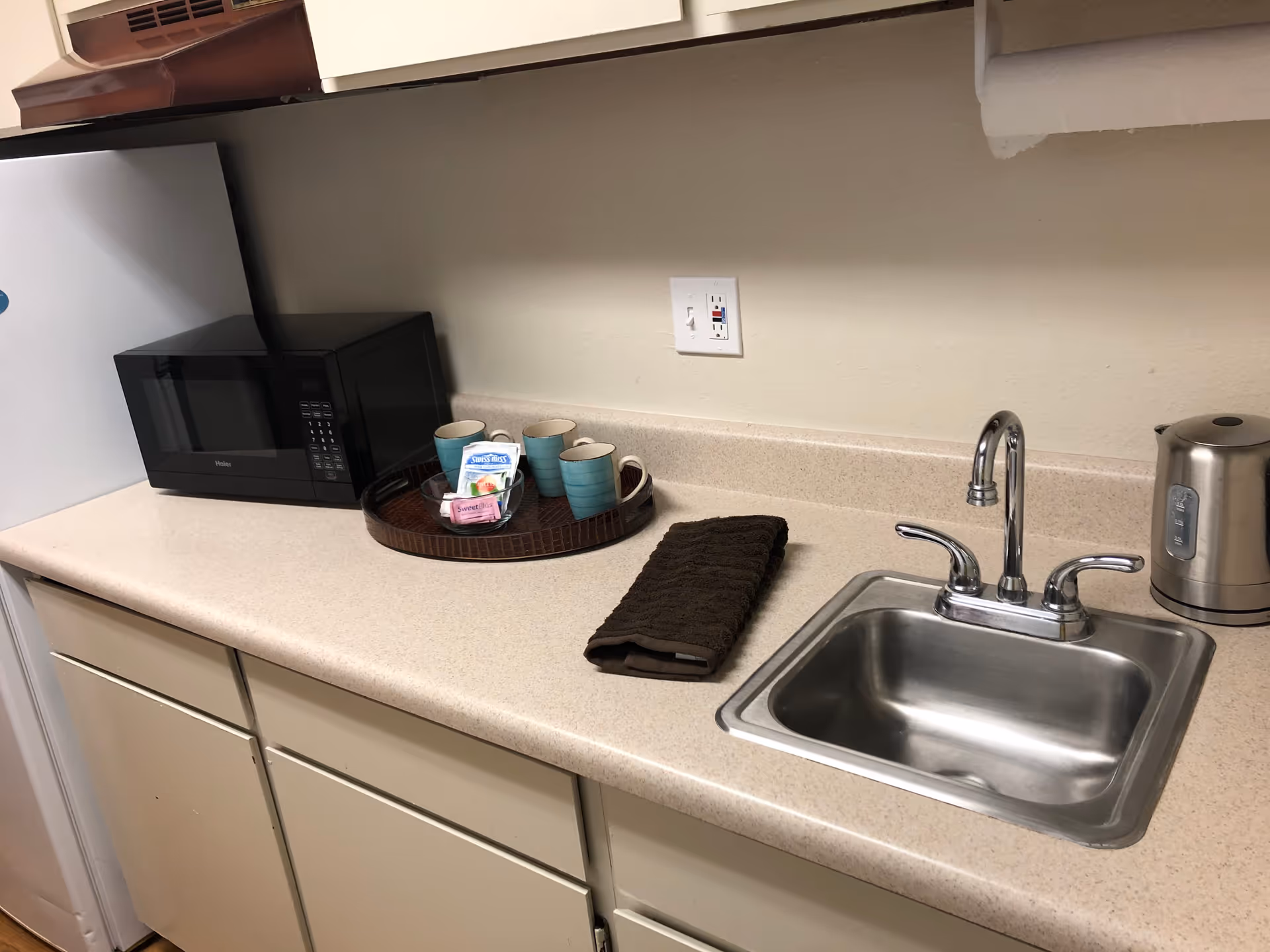 A kitchen countertop with a stainless steel sink, a silver electric kettle, a black microwave, and a round tray holding three blue mugs and packets of sweetener and creamer. A brown towel is folded next to the sink.