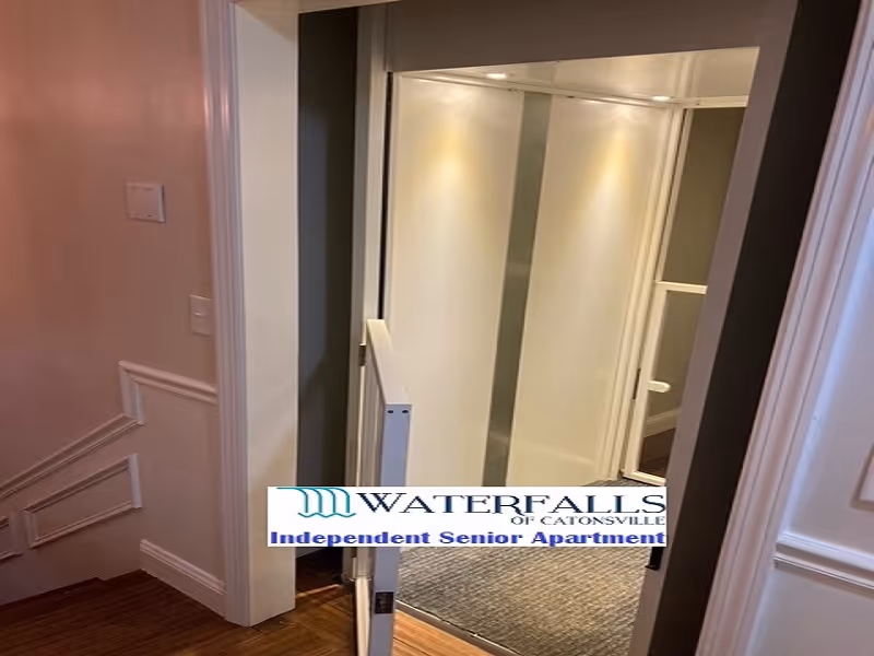 An interior view of a small residential elevator with its door open, showing a carpeted floor inside. The elevator is located next to a staircase with wooden steps and white walls with decorative molding.