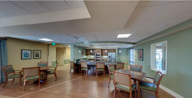 Interior view of a senior living community common area featuring a kitchen with a large island and bar stools, surrounded by multiple dining tables and chairs. The room has green walls, wooden flooring, framed artwork on the walls, and large windows letting in natural light.