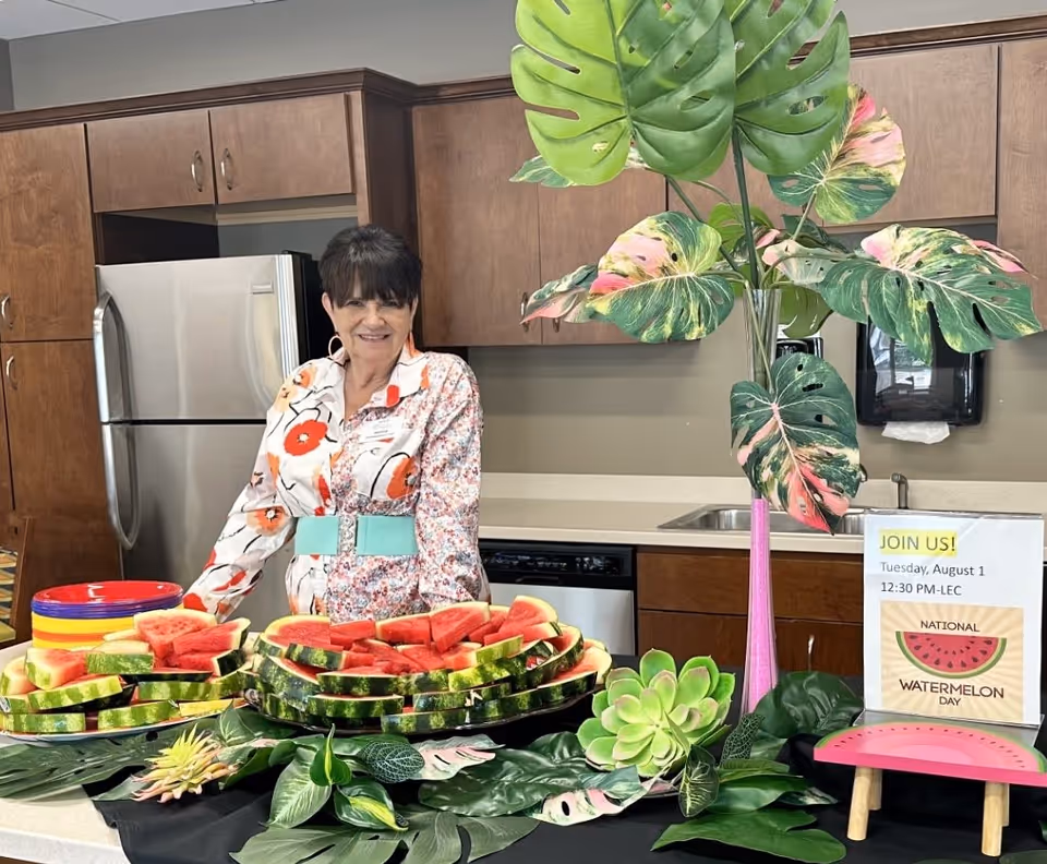 A woman stands behind a kitchen counter with plates of sliced watermelon arranged on it. The kitchen has wooden cabinets, a stainless steel refrigerator, and a sink. There is a tall vase with large green and pink leaves on the counter, along with a sign that reads 'JOIN US! Tuesday, August 1 12:30 PM-LEC NATIONAL WATERMELON DAY'.