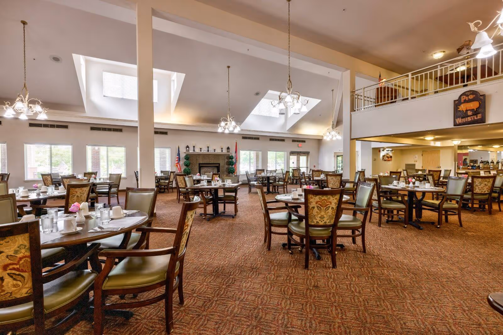 Spacious dining room with multiple round tables set with cups, glasses, and napkins. The room features high ceilings with skylights, chandeliers, large windows letting in natural light, and a fireplace at the far end. There is a second-floor balcony area and a sign that reads 'Pig & Whistle' on the wall.