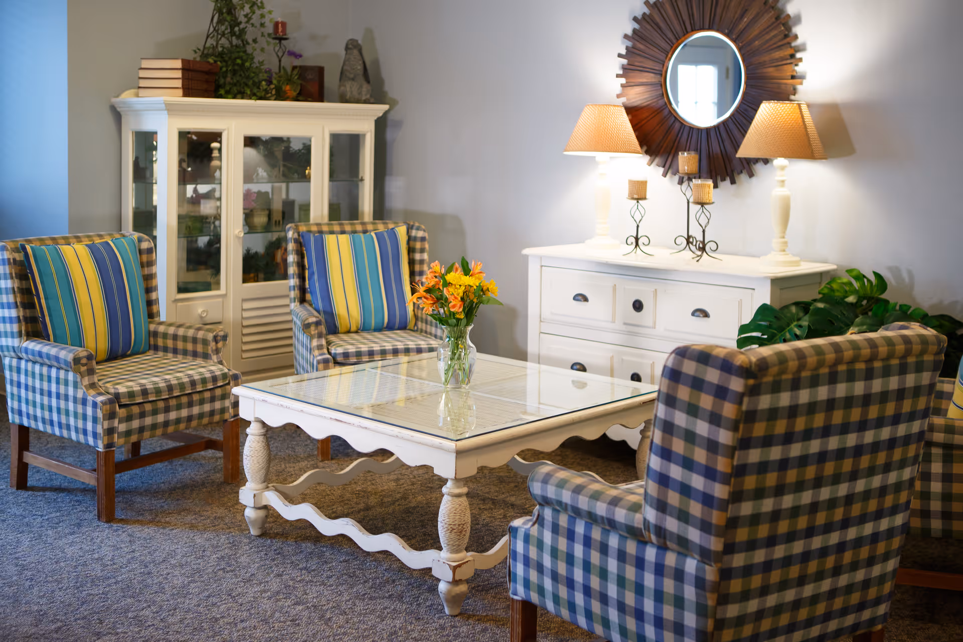A cozy sitting area with four plaid armchairs featuring blue, yellow, and green stripes, arranged around a white wooden coffee table with a glass top. On the table is a vase with yellow and orange flowers. Behind the chairs is a white cabinet with glass doors and decorative items on top, and a white dresser with two lamps, candles, and a round sunburst mirror hanging above it. The room has a carpeted floor and soft lighting.