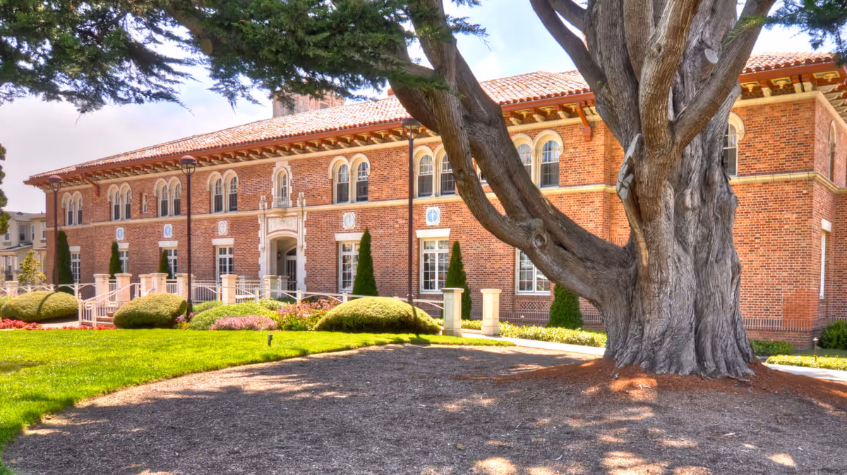 Exterior view of a large brick building with arched windows and a tiled roof, surrounded by well-maintained landscaping including bushes, grass, and a large tree in the foreground.