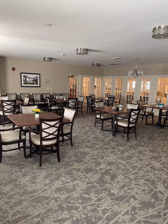 A spacious dining room with several wooden tables and cushioned chairs arranged neatly. The room has a patterned carpet, beige walls, and multiple ceiling lights. There are glass-paneled doors along one wall and a framed picture hanging on another wall. Small flower arrangements are placed on some tables.