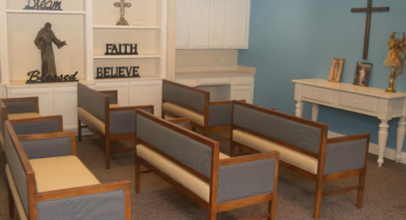 A small chapel or prayer room with several wooden benches with cushioned seats and backs arranged in rows. The walls are light blue and white, with religious decorations including a cross, a statue, and framed pictures on a white table. Shelves display words like 'Dream', 'Faith', 'Blessed', and 'Believe'.
