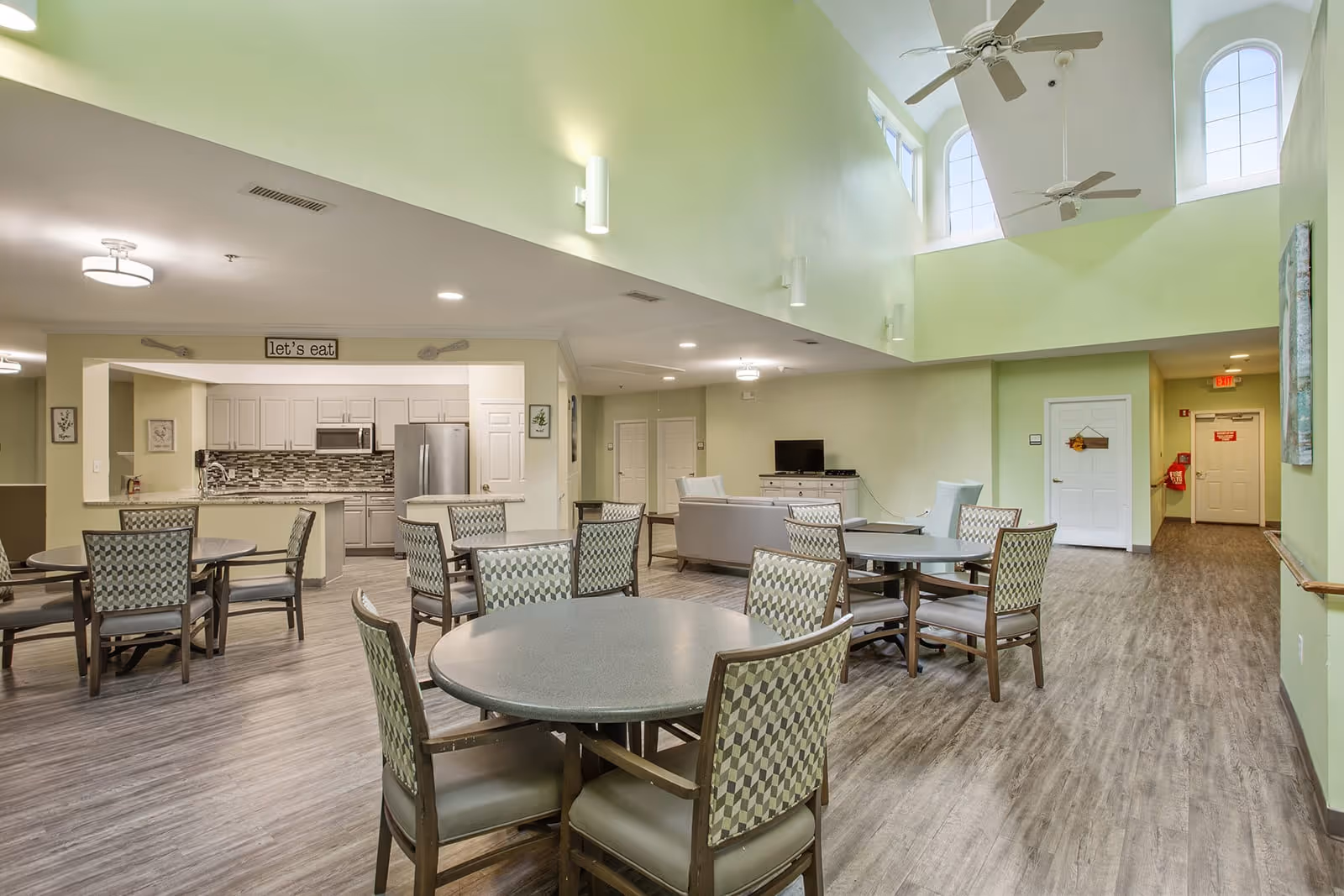 A spacious dining and living area in a senior living facility with round tables and chairs arranged for dining. The room features a high ceiling with ceiling fans and large windows near the ceiling. The kitchen area is visible in the background with white cabinets, a stainless steel refrigerator, and a sign above the kitchen opening that says 'let's eat'. There is a seating area with sofas and a TV on a stand. The flooring is wood-style laminate, and the walls are painted light green.