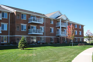 Exterior view of a three-story brick apartment building with balconies, surrounded by a well-maintained lawn, small trees, and a sidewalk under a clear blue sky.