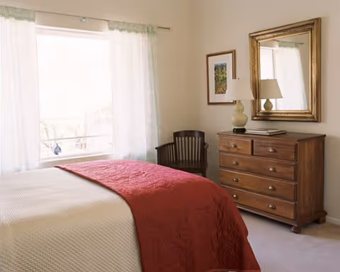 A bedroom with a bed dressed in white and red linens, a wooden dresser with a lamp and mirror, a chair, and a window with sheer curtains.