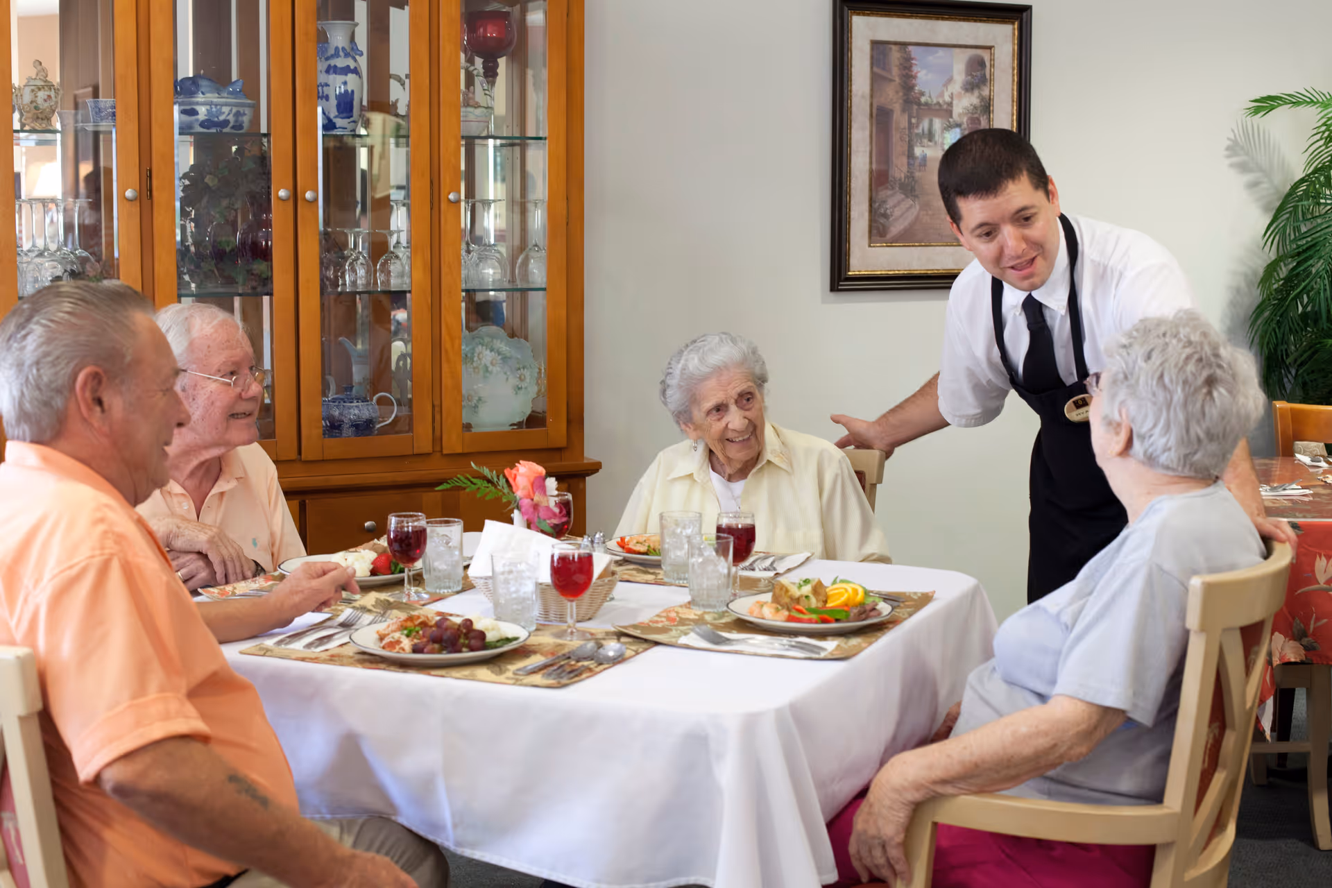 Four elderly people seated around a dining table with plates of food and glasses of red drink, while a waiter in a white shirt and black apron leans in to speak with them in a dining room setting with a wooden cabinet and framed artwork on the wall.
