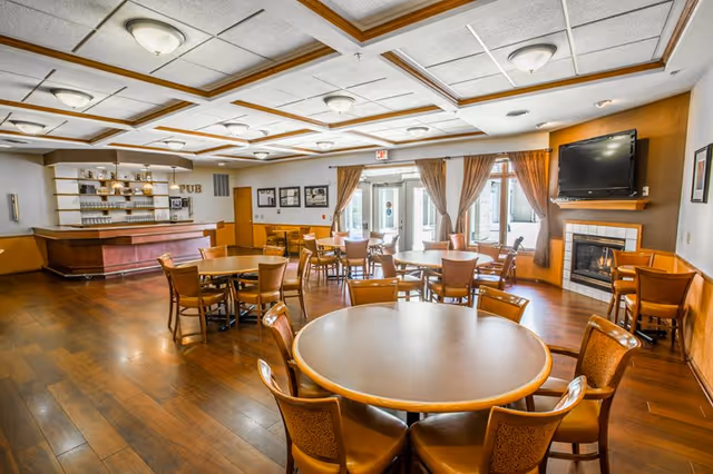 A spacious common area with multiple round wooden tables and chairs arranged on a wooden floor. There is a fireplace with a mounted TV above it on the right side, and a bar area labeled 'PUB' on the left side. Large windows with brown curtains allow natural light to enter the room.