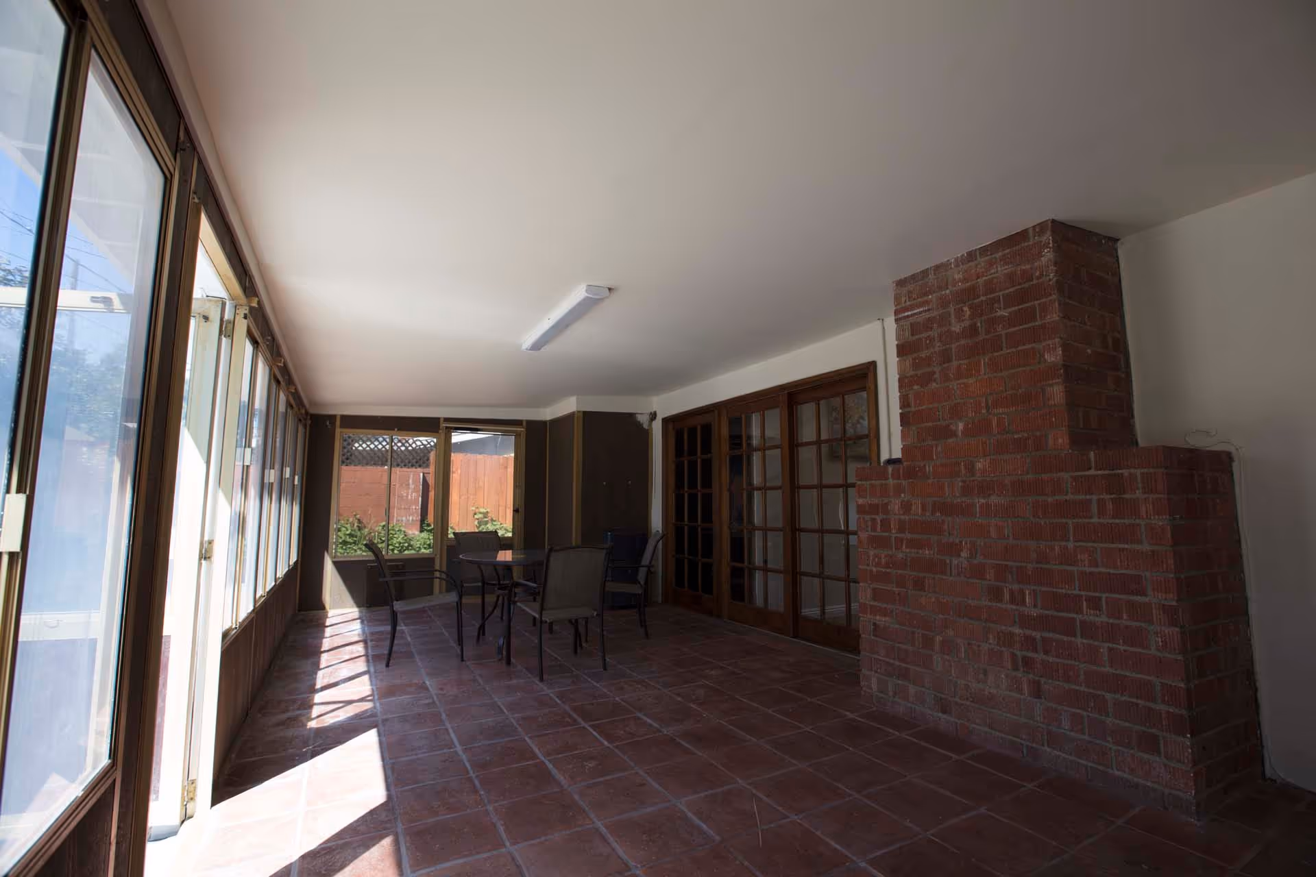 A sunlit enclosed patio area with large windows on one side, a brick fireplace on the right, and a set of glass doors leading to another room. There is a small table with four chairs in the middle of the space, and the floor is covered with reddish-brown tiles.