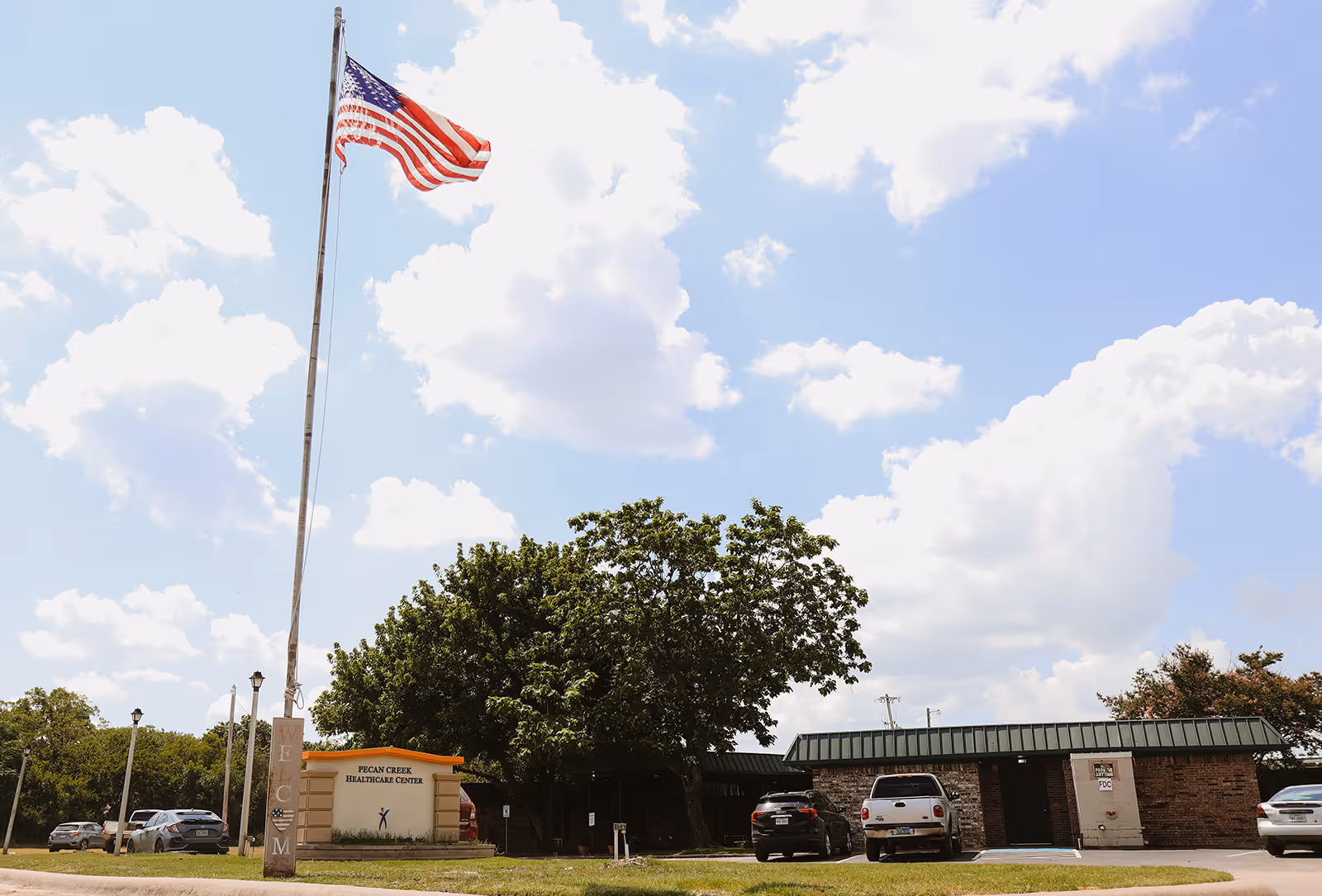 Front exterior of Pecan Creek Healthcare Center with a tall flagpole flying an American flag, the facility sign, trees, and parked cars under a blue sky.
