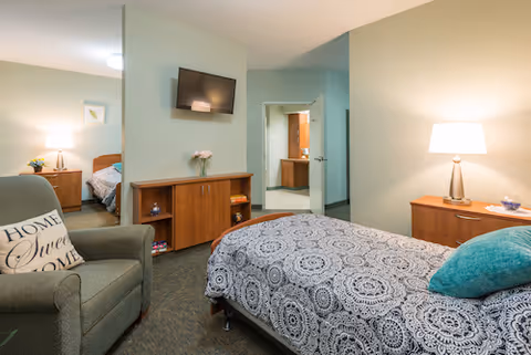 A cozy bedroom in a senior living facility featuring a bed with a patterned bedspread and a teal pillow, a wooden dresser with a lamp, a comfortable armchair with a 'Home Sweet Home' pillow, a wall-mounted flat-screen TV, and an open doorway leading to a bathroom and another bedroom.