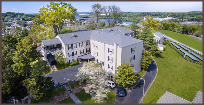 Aerial view of a multi-story senior living facility building surrounded by trees and greenery, with a parking area and a driveway. The building is light-colored with multiple windows and a covered entrance. In the background, there is a river and a bridge, along with a small town and more trees.