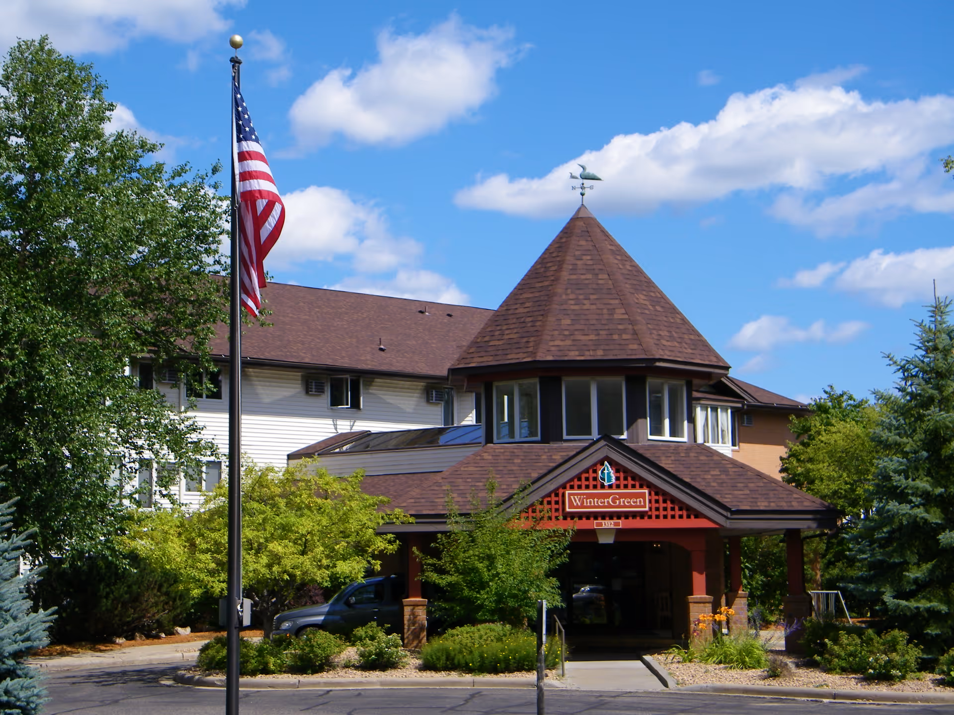 Exterior view of a senior living facility building with a peaked roof and multiple windows. The entrance has a sign that reads 'WinterGreen' and is surrounded by trees and shrubs. An American flag is flying on a flagpole in front of the building under a blue sky with scattered clouds.