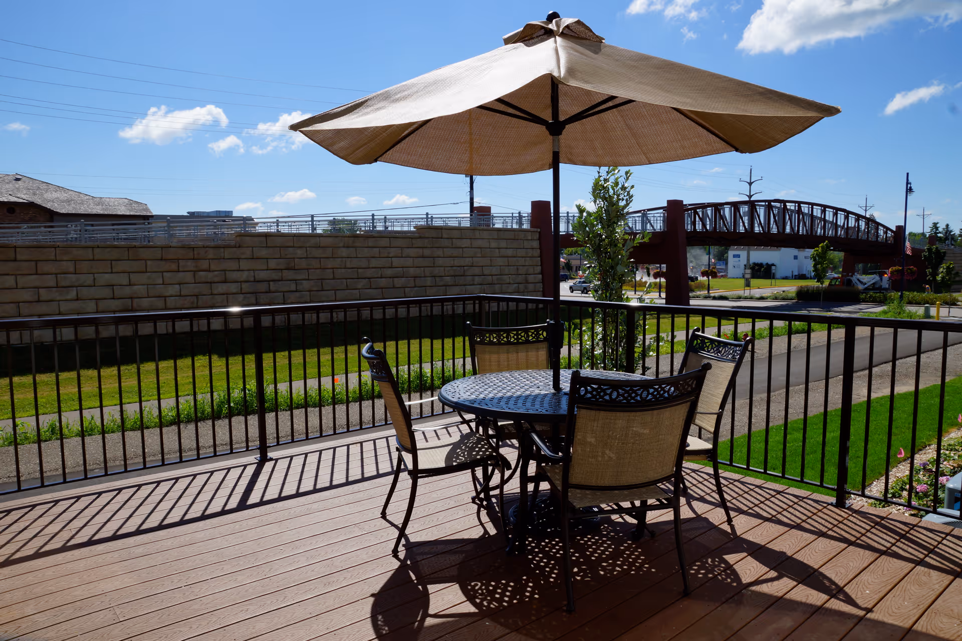 Outdoor patio area with a round metal table and four chairs under a large beige umbrella. The patio has a wooden floor and black metal railing. In the background, there is a stone wall, a pedestrian bridge, green grass, and a clear blue sky with a few clouds.