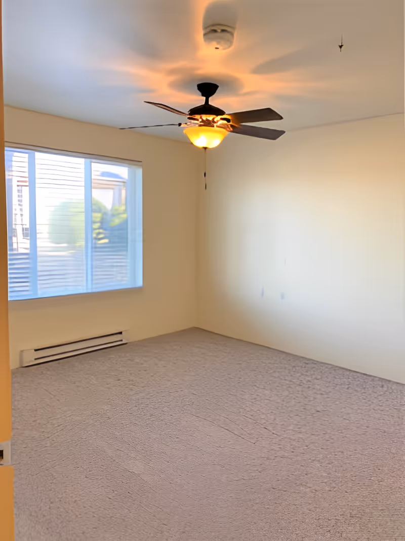 Empty room with beige carpet, cream-colored walls, a ceiling fan with light, and a large window with blinds letting in natural light.