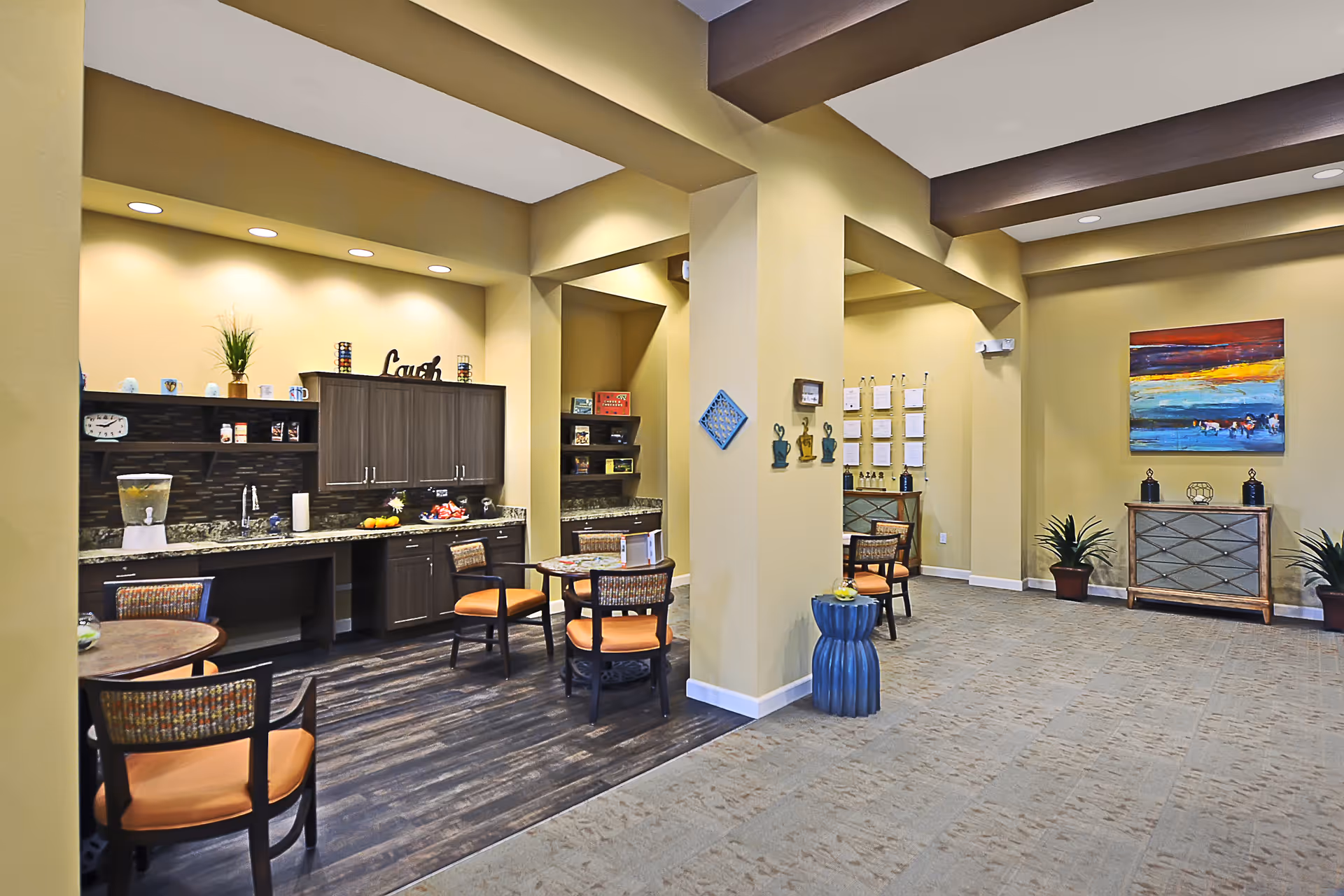 A bright and spacious common area in a senior living facility featuring a small kitchenette with dark cabinets, a granite countertop, and a water dispenser. There are round tables with chairs that have orange cushions. The walls are painted yellow, and there is colorful artwork and decorative plants placed around the room.