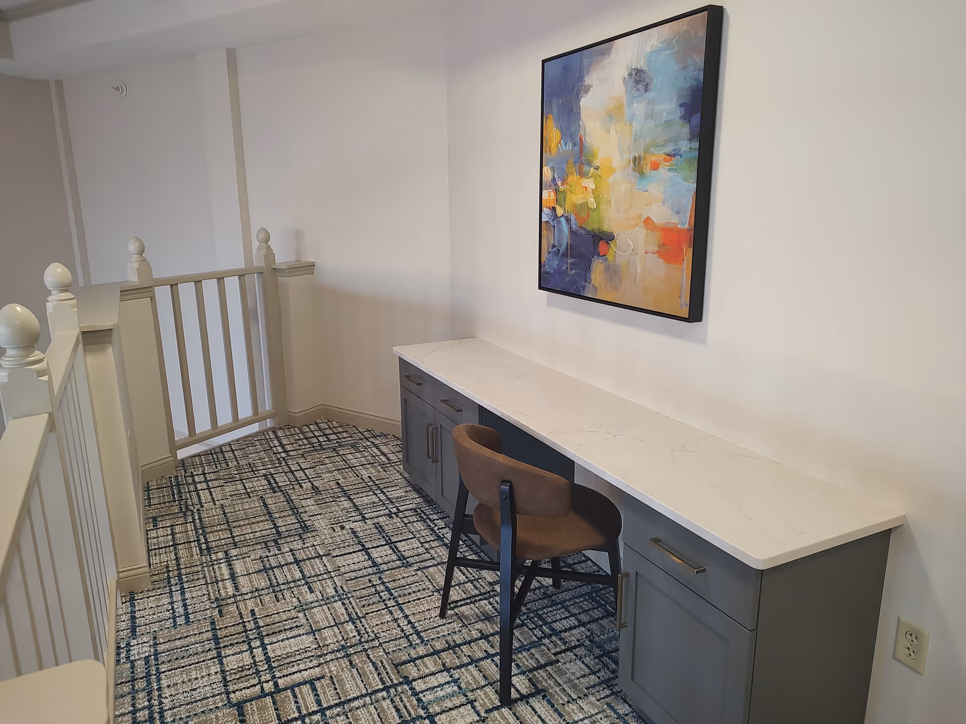 A small workspace area with a long white countertop and gray cabinets underneath. A brown chair is placed in front of the counter. Above the counter hangs a colorful abstract painting. The floor is carpeted with a patterned design in shades of blue and beige. A white railing is visible on the left side.