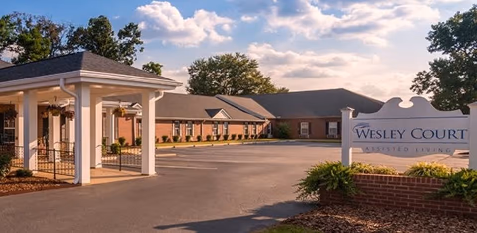Exterior view of Wesley Court assisted living facility showing a brick building with a covered entrance, parking area, and a sign with the facility name surrounded by greenery under a partly cloudy sky.