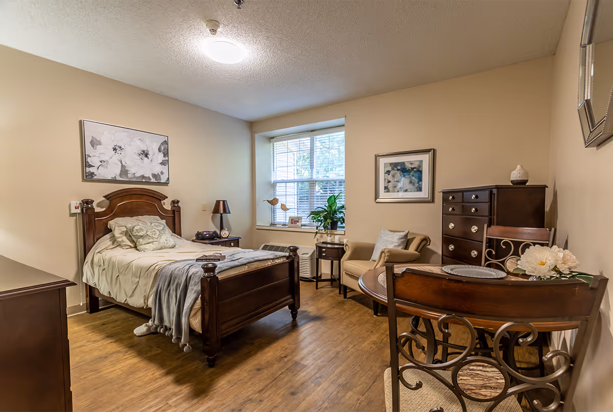A cozy bedroom in a senior living facility featuring a wooden bed with beige bedding and pillows, a nightstand with a lamp and clock, a window with blinds, a small armchair with a pillow, a wooden dresser, a round wooden table with two chairs, and framed floral artwork on the walls.