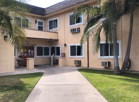 Front entrance of a two-story beige building with a walkway, palm trees, windows and window air conditioners.