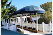 Covered walkway with a blue canopy and white picket fences on both sides, leading to a building surrounded by trees and greenery.