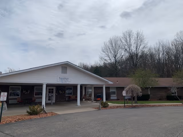 Exterior front view of a single-story senior living facility building with a covered entrance supported by white columns. There are some chairs on the porch, a small tree with white blossoms, and leafless trees in the background under a cloudy sky.