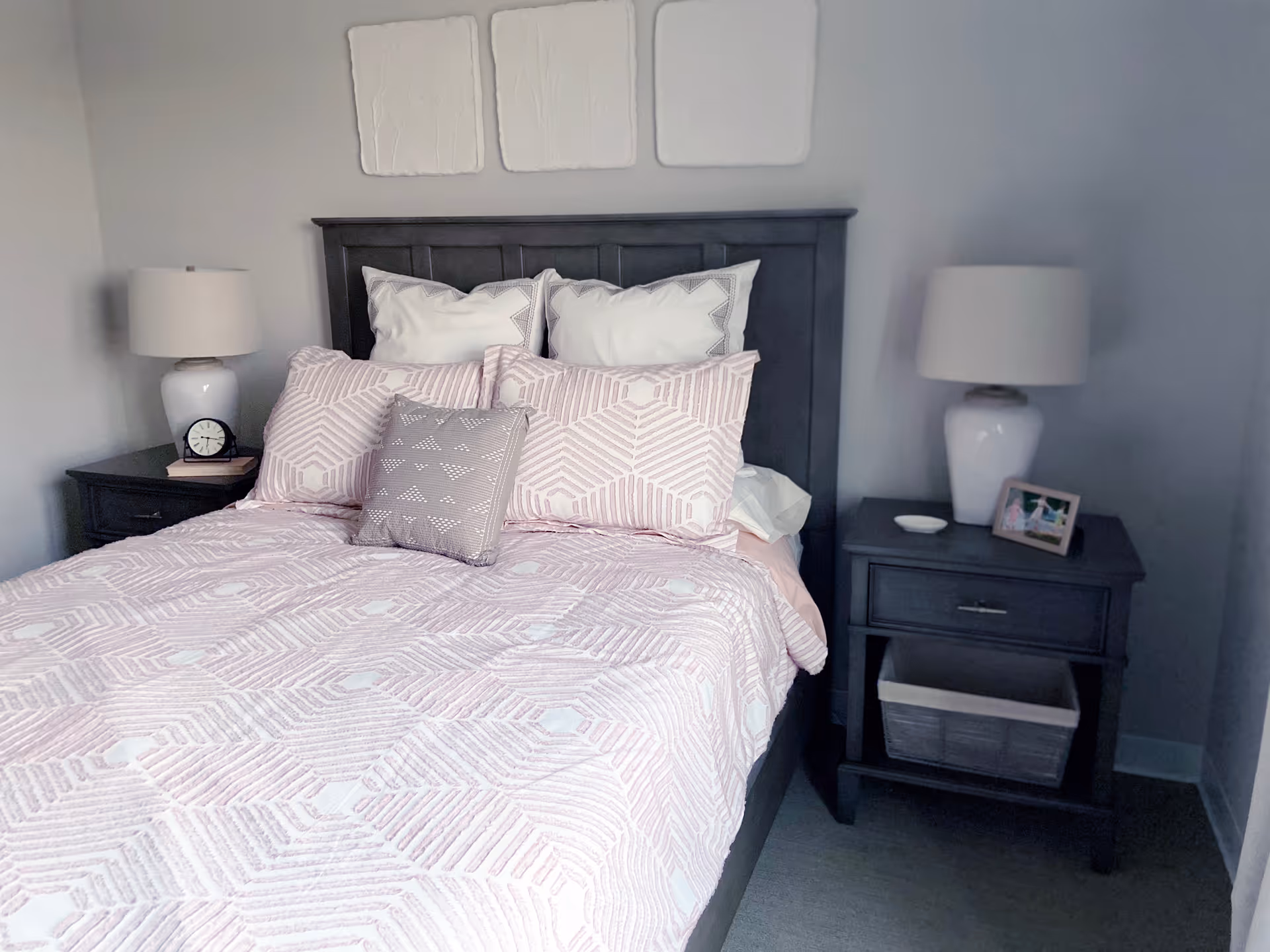 A neatly made bed with a dark wooden headboard, adorned with multiple pillows in white and light pink geometric patterns. On each side of the bed are matching dark wooden nightstands, each with a white lamp. One nightstand has a small clock and a book, while the other has a framed photo and a small dish. The walls are painted light gray with three white decorative panels hanging above the bed.