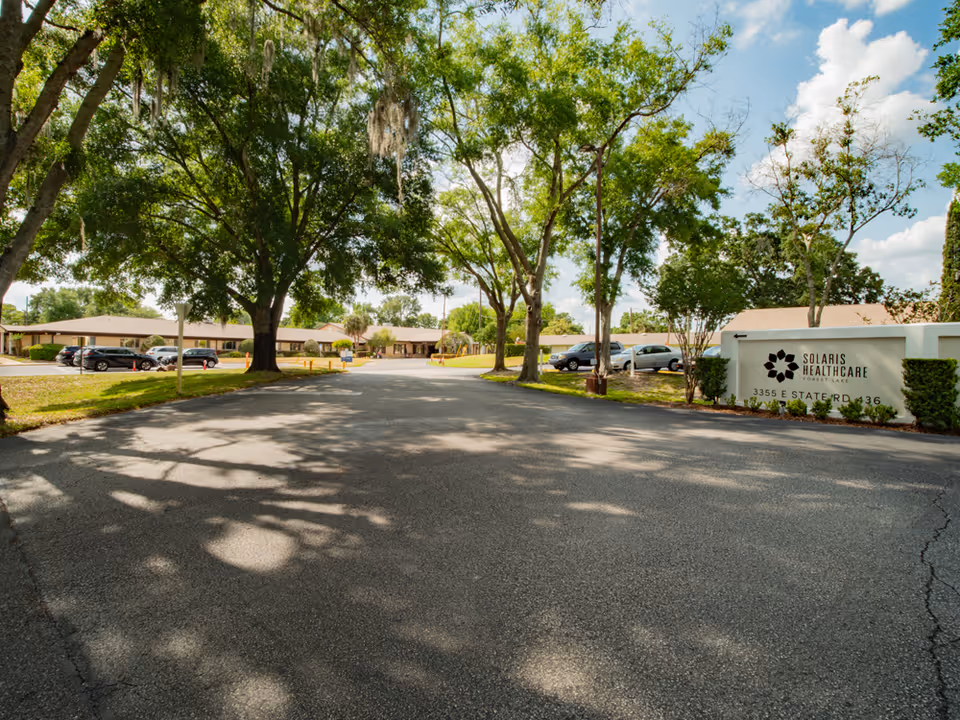 Entrance driveway to Solaris Healthcare Forest Lake facility with large trees lining the road and a sign displaying the facility name and address on the right side. Several parked cars are visible near the single-story building under a partly cloudy sky.