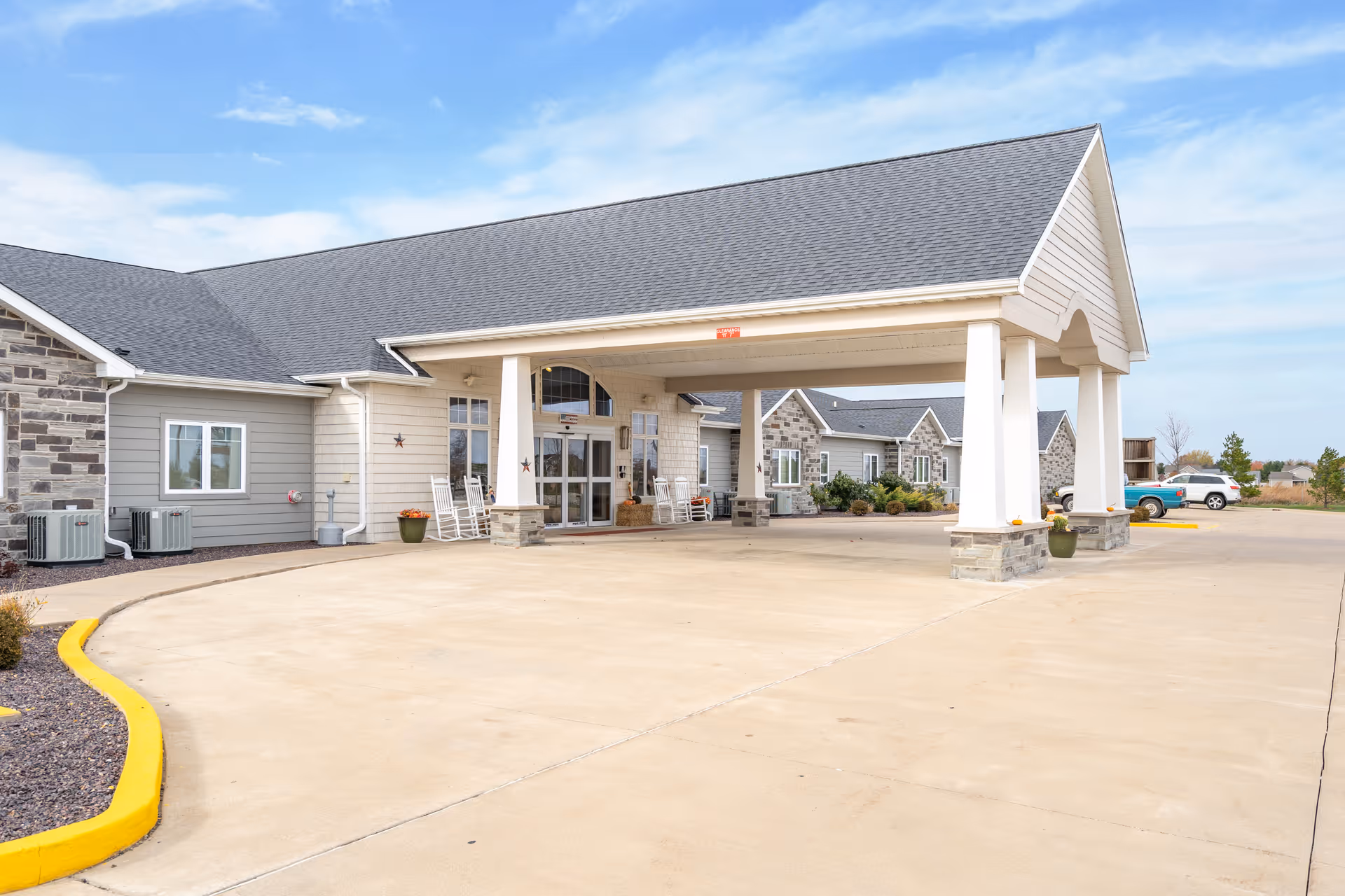 Exterior view of Reflections Memory Care facility in Washington, IL, showing a covered entrance with white pillars, rocking chairs, potted plants, and a spacious driveway under a partly cloudy sky.