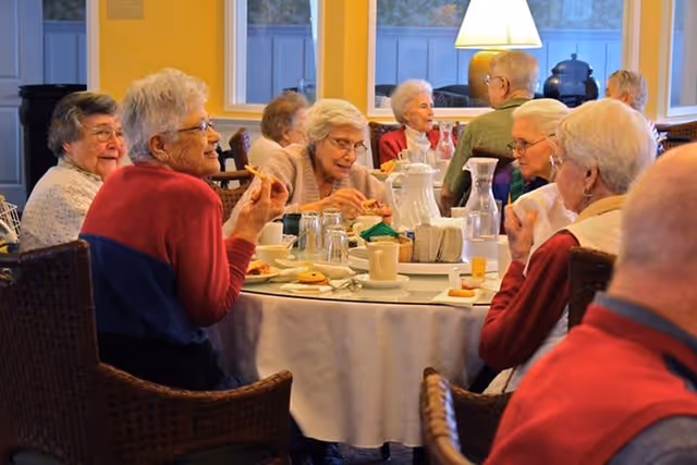 Several elderly residents seated around a round table in a bright dining room, eating and conversing.