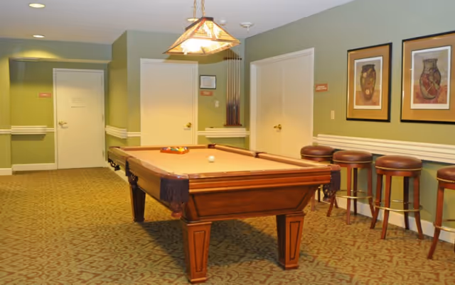 A recreational room with a pool table in the center, a hanging light fixture above it, four bar stools lined up against the wall, and framed artwork on the wall. The room has green walls, patterned carpet, and three closed doors.