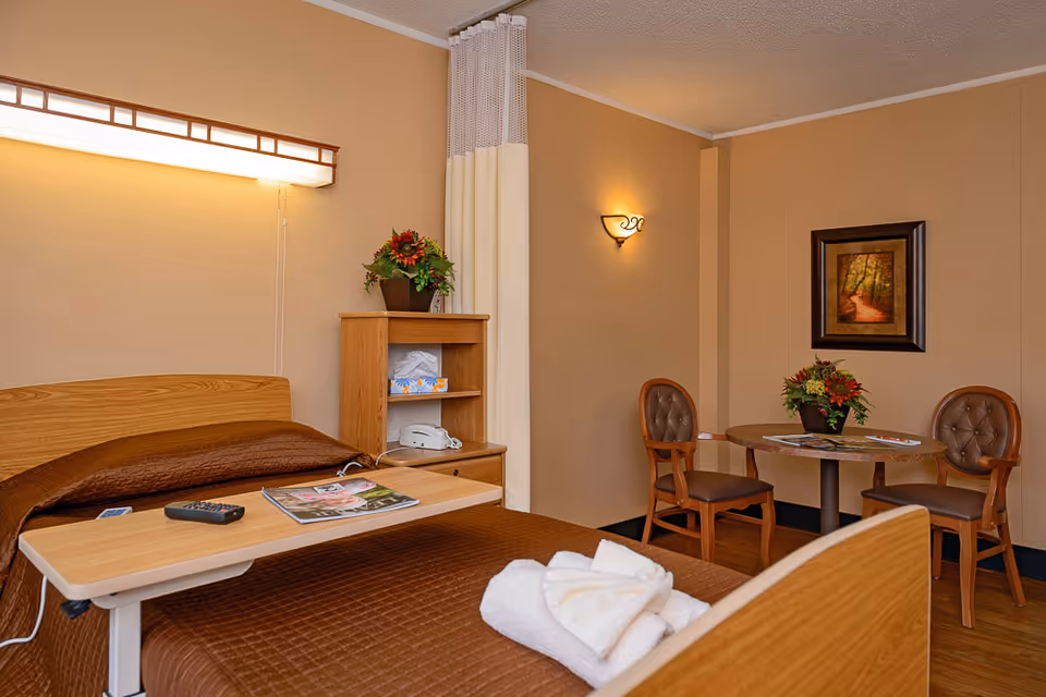 A cozy senior living bedroom with a bed and overbed table, a small round table with two chairs, shelving, plants, and wall art.