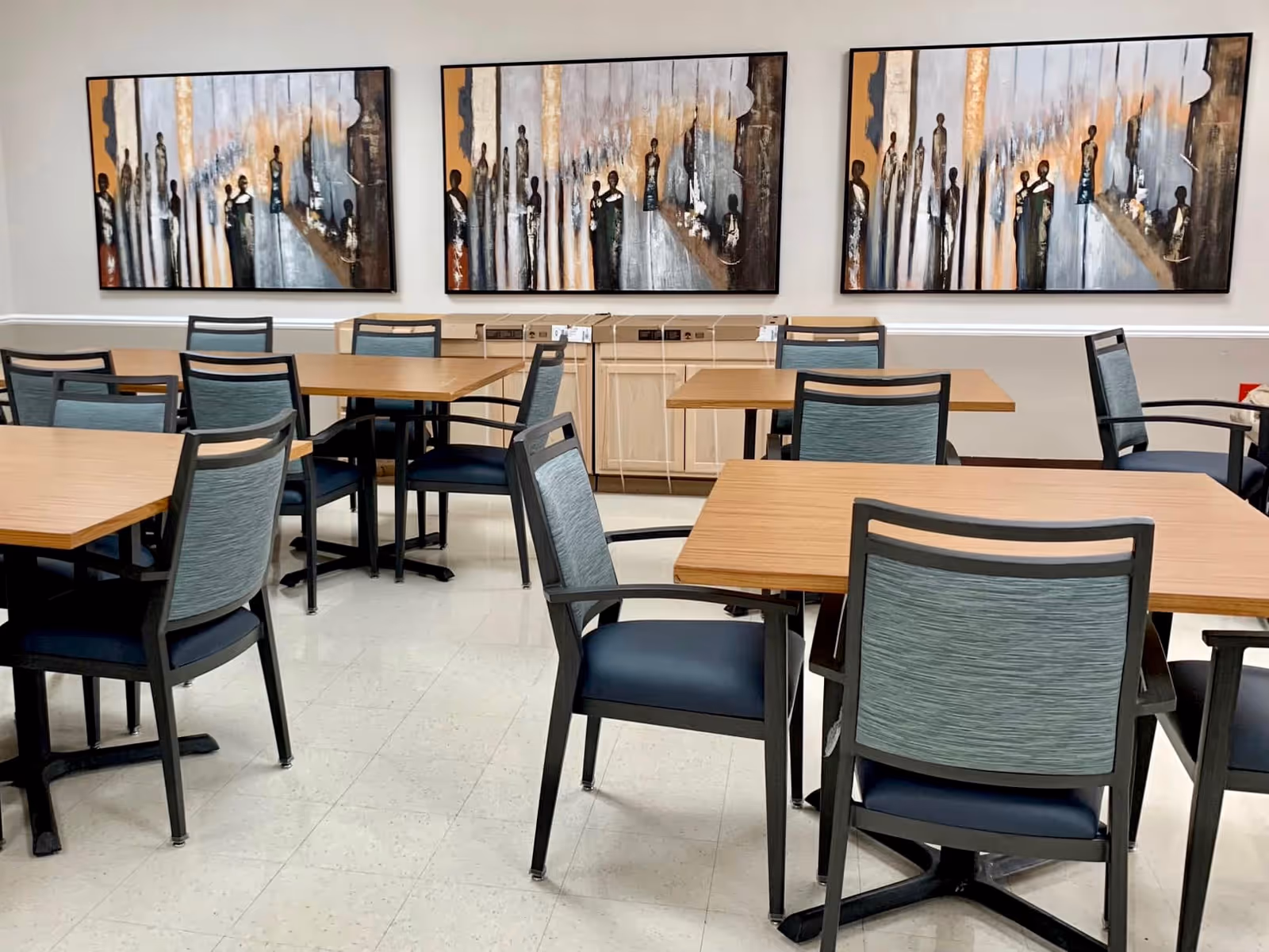 A dining area with multiple wooden tables and chairs with blue cushions. Three abstract paintings featuring elongated human figures are hung on the wall above a light-colored cabinet.