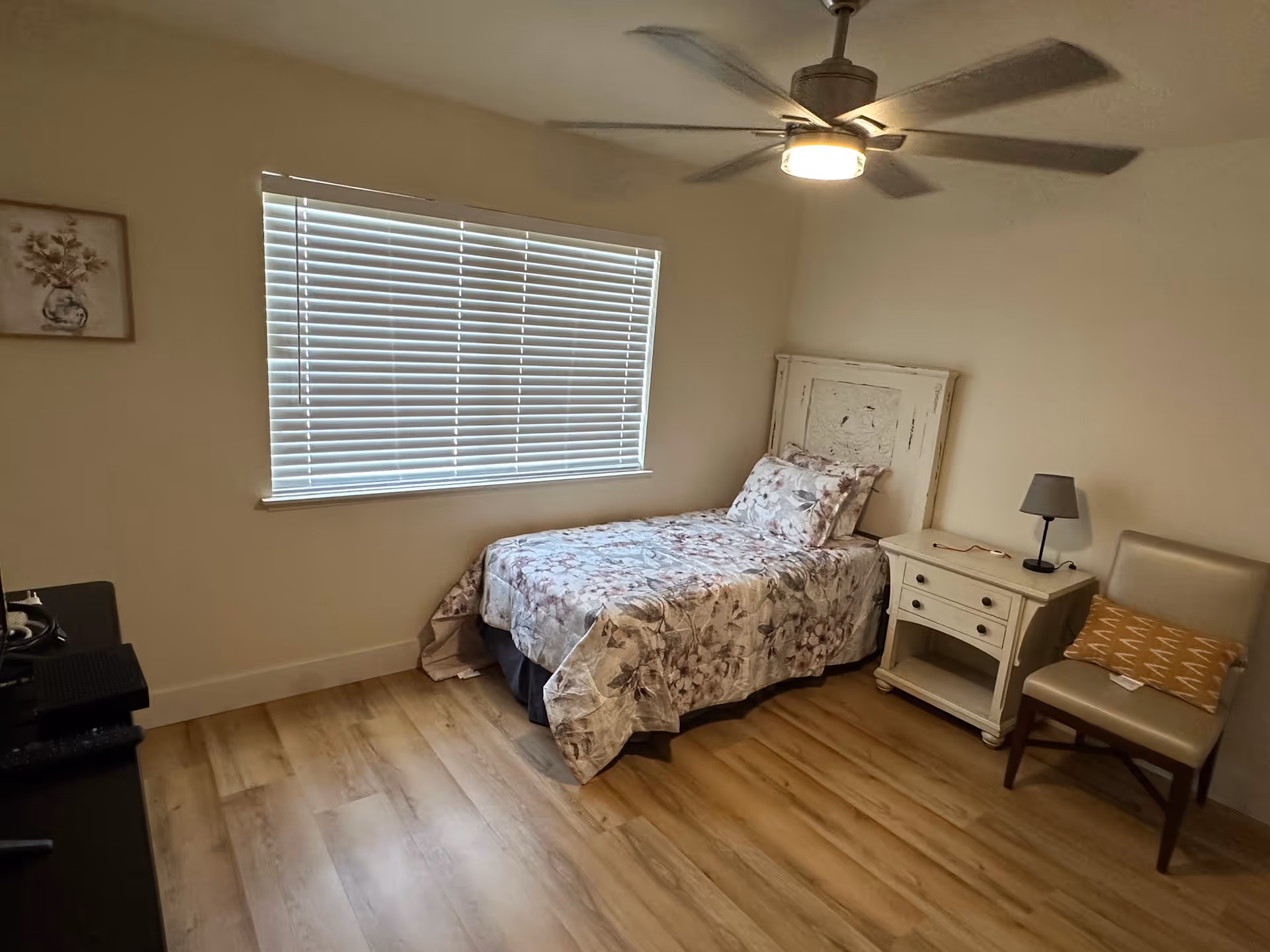A small bedroom with a single bed covered in floral bedding, a white nightstand with a small lamp, a beige chair with a patterned cushion, a large window with closed blinds, and a ceiling fan with a light.