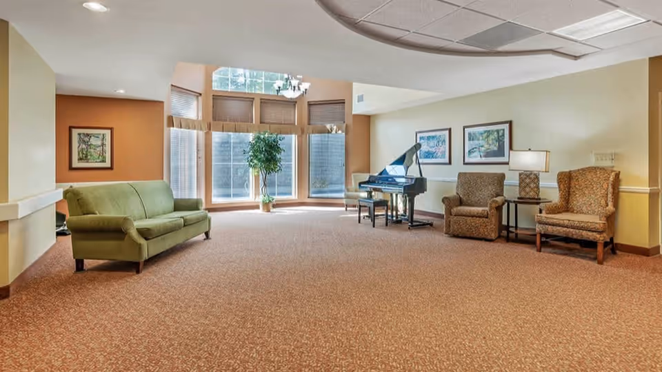 A spacious living room area with a green sofa on the left, two patterned armchairs on the right separated by a small table with a lamp, a black grand piano near large windows with blinds, and a potted plant in front of the windows. The room has beige walls, carpeted floor, and framed pictures on the walls.