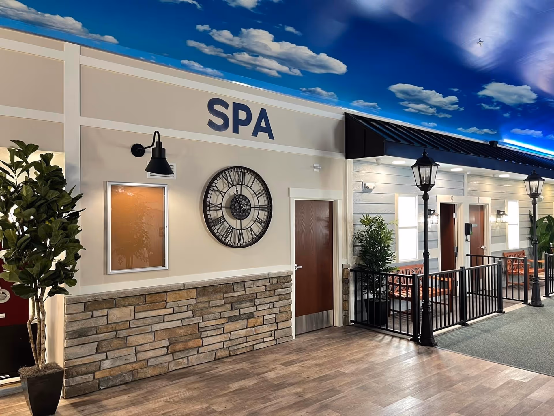 Indoor hallway area with a spa entrance marked by a large clock on the wall and the word 'SPA' above the door. The ceiling is painted with a blue sky and white clouds. There are potted plants, black metal railings, streetlamp-style lights, and wooden benches along the corridor.