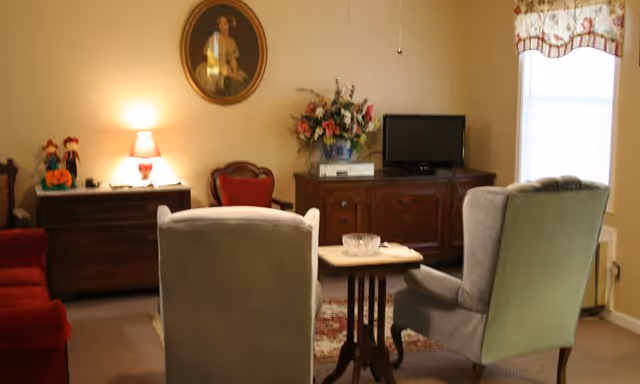 A cozy living room with two gray armchairs facing a small wooden table. Behind the table is a wooden dresser with a flat-screen TV and a large floral arrangement. To the left, there is another wooden dresser with a lamp, decorative items, and a framed portrait hanging on the wall above it. A window with a floral valance is on the right side of the room.