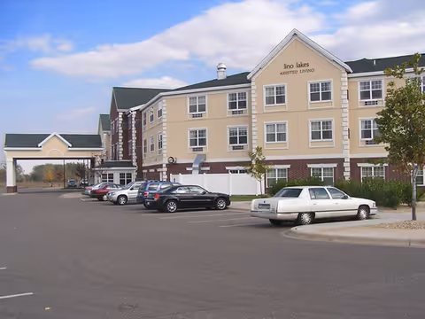 Three-story Lino Lakes Assisted Living building with a covered entrance and parked cars in the foreground.