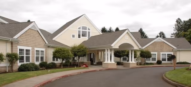 Exterior front of a beige senior living building with a covered entrance porte-cochère, driveway, and landscaped shrubs.