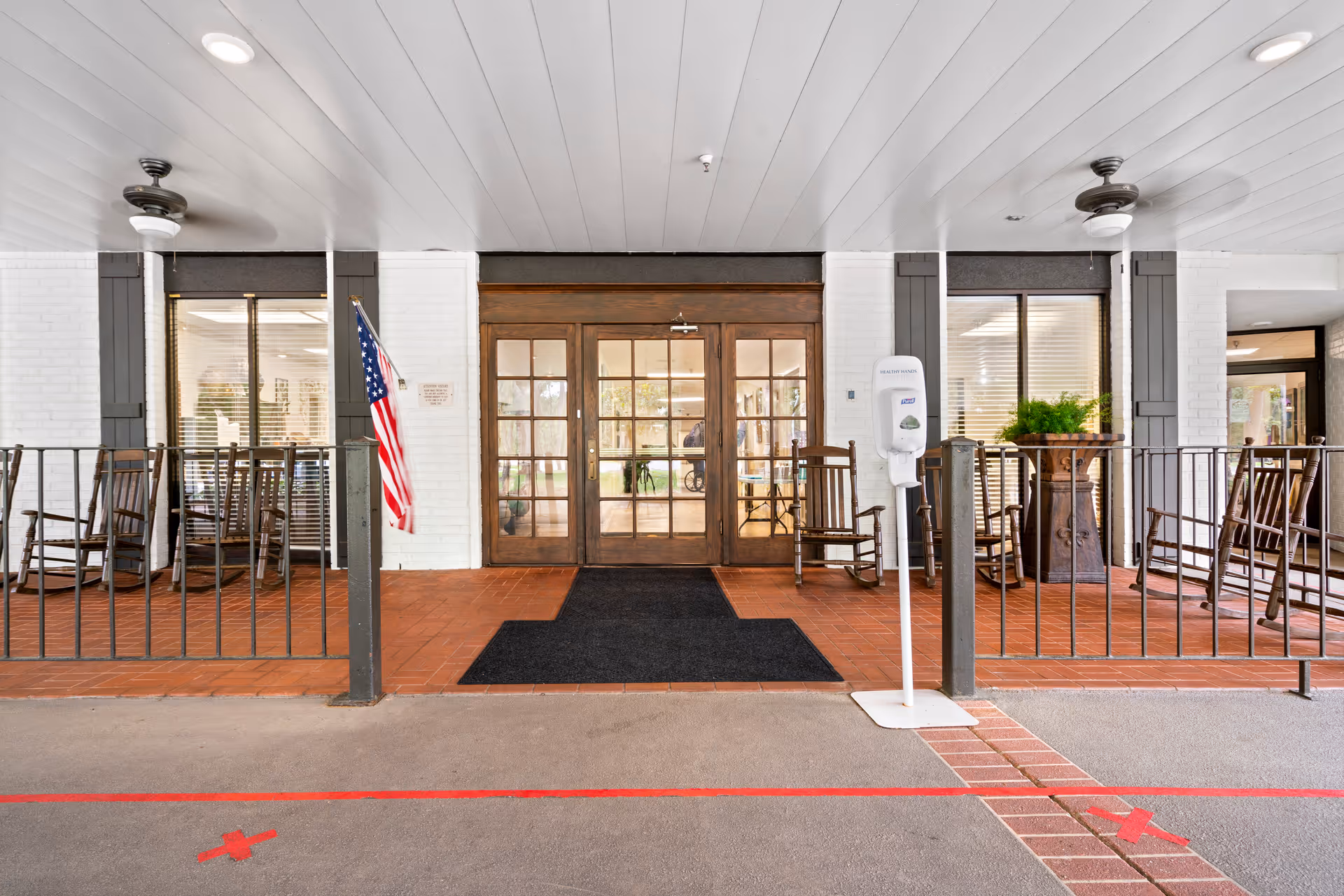 Entrance area of Heritage Manor of Mandeville featuring double wooden glass doors, an American flag mounted on the left side, several wooden rocking chairs on both sides of the entrance, a hand sanitizer dispenser on a stand to the right, and ceiling fans above. The floor is tiled near the entrance with a black mat leading to the doors, and the area outside has a red line marked on the ground.